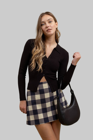 Woman wearing a black cardigan and plaid skirt, holding a black handbag against a green background