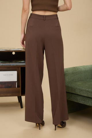 Person wearing brown high-waisted pants standing in a room with a beige wall and green sofa.