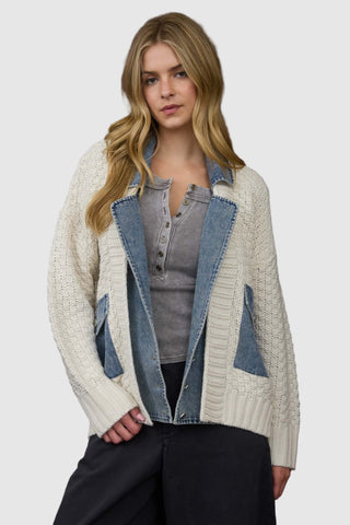 Woman wearing a white cardigan with denim accents over a gray shirt against a wooden paneled wall.