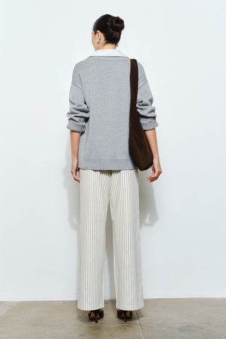 Person wearing a gray sweater and white pants with a brown bag, standing against a white wall.