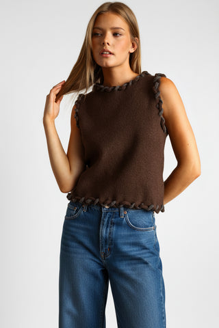 A woman modeling a sleeveless brown knit mini dress with dark brown braided trim details at the neckline, waist, and hem, set against a solid warm brown studio background.