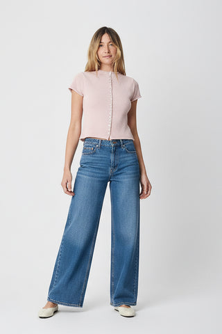 A woman modeling a light pink two-piece lounge set consisting of a short-sleeve button-front top and matching drawstring shorts, paired with cream-colored flats, against a light gray studio background.