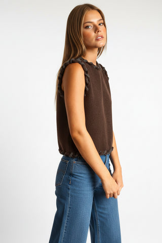 A woman modeling a sleeveless dark brown knit dress with ruffled trim, posed against a solid warm tan studio background.