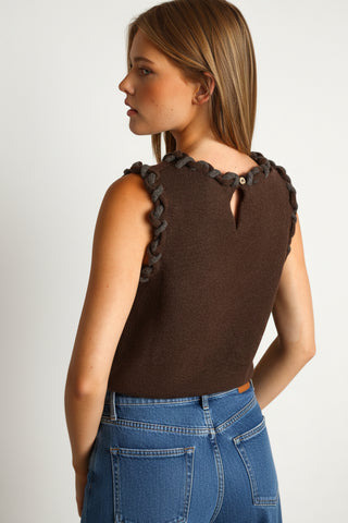 A woman shown from the back wearing a sleeveless dark brown knit top with a dark gray braided trim along the shoulder straps and a small button detail at the back neckline, against a warm tan background.