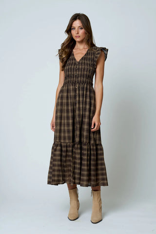 A woman modeling a brown and black plaid midi dress with ruffled cap sleeves, a V-neckline, and a tiered skirt, paired with beige ankle boots, against a light gray studio background.