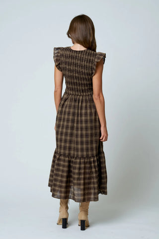 A woman viewed from the back wearing a brown and beige plaid midi dress with ruffled cap sleeves and a tiered skirt, paired with beige ankle boots, against a light gray studio background.