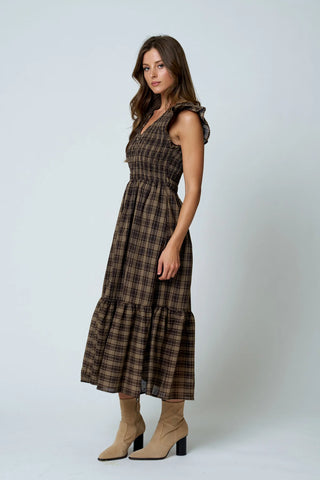 A woman modeling a sleeveless brown and beige plaid midi dress with ruffled shoulder straps and a tiered skirt, paired with beige ankle boots, against a light gray studio background.