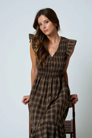 A woman wearing a brown and beige plaid midi dress with ruffled cap sleeves and a smocked waist, standing next to a wooden chair against a light gray background.