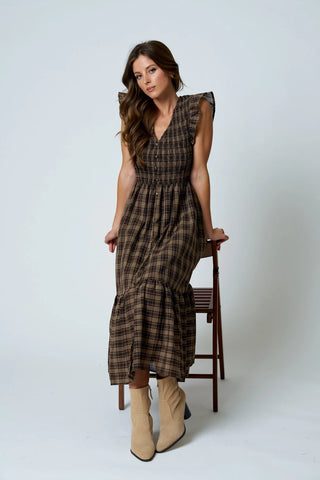 A woman models a brown and beige plaid midi dress with ruffled cap sleeves and a tiered skirt, standing next to a wooden chair against a light gray studio background.