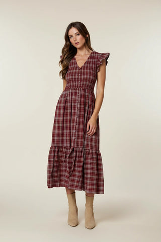 A woman wearing a red and dark plaid midi dress with ruffle sleeves and beige ankle boots, standing in a lush garden greenhouse setting.
