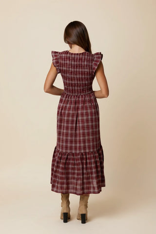 A woman viewed from behind wearing a red and dark plaid midi dress with ruffled cap sleeves and a tiered hem, standing in a greenhouse surrounded by potted plants.