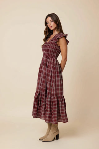 A woman wearing a red and dark plaid midi dress with ruffled cap sleeves and beige ankle boots, standing in a greenhouse surrounded by potted plants.