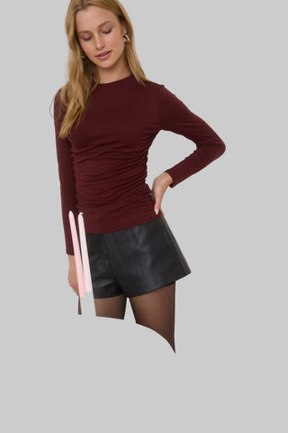 Woman wearing a burgundy long-sleeve top and black leather shorts on a gray background