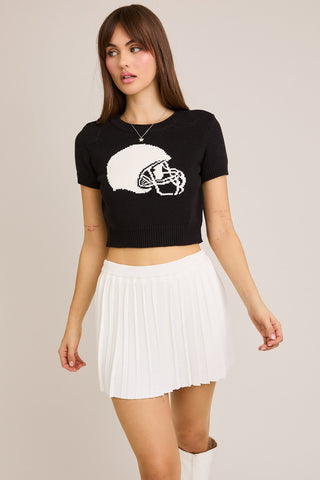 Woman wearing a black crop top with a helmet design and a white pleated skirt on a beige background