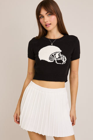 Woman wearing a black sweater with a white football helmet graphic design and a white pleated skirt on a beige background