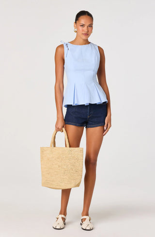 Woman wearing a light blue sleeveless top and dark denim shorts, holding a straw bag on a white background