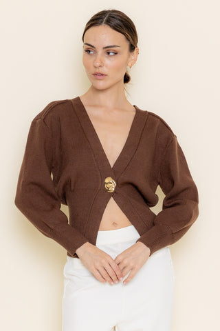 Woman wearing a brown cardigan with a gold emblem on a beige background