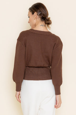 Woman wearing a brown sweater and white pants against a beige background