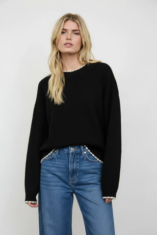A blonde model wearing a black knit sweater with white ruffle trim at the collar, hem, and cuffs, paired with white ruffled shorts, against a light gray studio background.