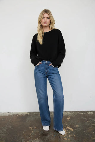 A woman stands against a white wall wearing a black crewneck sweater, white frayed shorts, and white knee-high socks.
