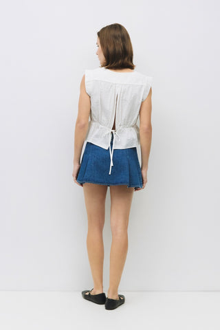 Back view of a white textured sleeveless top with a center slit and adjustable tie at the waist, styled with a pleated denim mini skirt