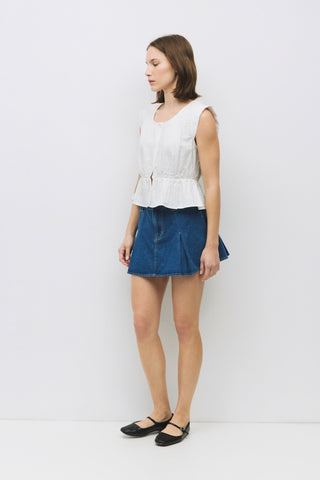 Side profile of a woman wearing a sleeveless white peplum top with a button front and cinched waist, styled with a denim mini skirt and black flats