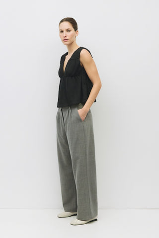 Side angle of sleeveless black V-neck top with gathered waist, paired with gray pleated wide-leg trousers and ivory flats