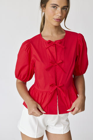 Close-up front view of red puff sleeve top with three bow tie closures, styled with white shorts.