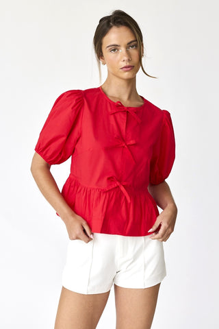 Model wearing red puff sleeve cotton blouse with three front bow ties, paired with white shorts.
