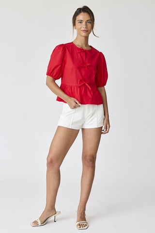 Full-body view of model wearing red puff sleeve bow tie top with white shorts and heeled sandals.