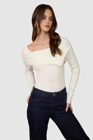 Woman wearing a white off-shoulder top and dark blue jeans against a red curtain background.