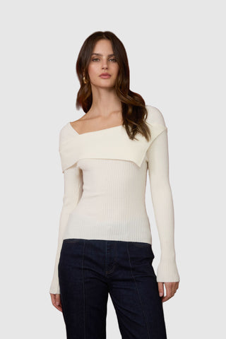 Woman wearing a white off-shoulder sweater and dark pants against a red curtain background