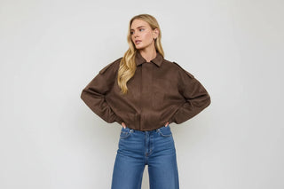 A blonde model wearing a brown suede-like cropped jacket with a collar and front pockets, paired with a white cow-print mini skirt, against a plain light gray studio background.
