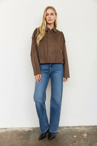 A woman modeling a brown suede cropped jacket and a white cow-print mini skirt, paired with black knee-high boots, standing against a plain white wall.
