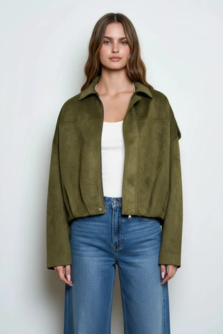 A woman modeling an olive green cropped suede jacket over a white top and a dark brown ruffled tiered skirt, photographed against a plain light gray studio background.