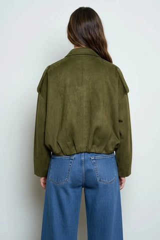 A woman viewed from behind wearing an olive green suede jacket and a dark brown ruffled mini skirt, set against a plain light gray background.