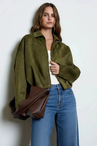 A woman models an oversized olive green suede jacket, styled with a white top, a brown ruffled mini skirt, and a large dark brown shoulder bag against a plain light gray background.