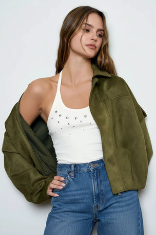 A woman modeling an olive green suede jacket worn open over a white studded tank top and dark brown ruffled pants, set against a plain light gray studio background.