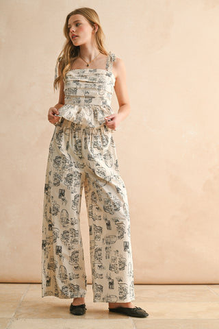 Model wearing a cream ruffled peplum top with black illustrated print and bow tie straps, paired with matching wide-leg pants and black ballet flats.