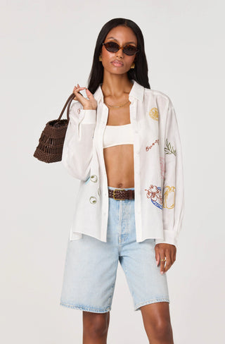 Woman wearing a white embroidered jacket, white top, and light blue shorts with a brown bag.