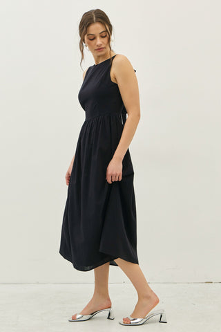 Model in a black sleeveless cotton midi dress shown in angled side view with gathered waist and flowy skirt