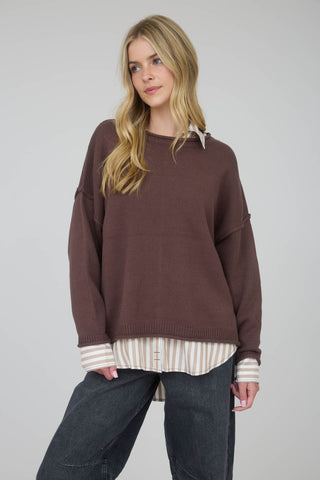 Woman wearing a brown sweater over a striped shirt with dark jeans on a plain background