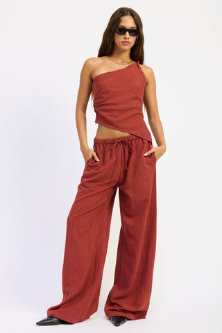 A woman modeling a rust-red two-piece set consisting of a one-shoulder wrap top and matching wide-leg pants with vertical pinstripes, set against a plain white studio background.