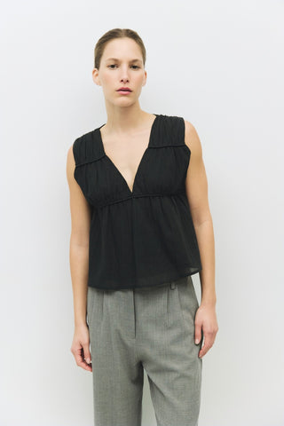 Model wearing a sleeveless black cotton top with deep V-neckline and gathered waist, styled with gray pleated trousers