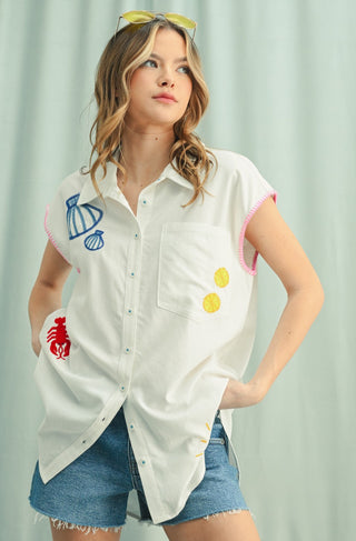 Front view of sleeveless white button-up tunic with colorful embroidered summer motifs and pink trim, styled with denim shorts