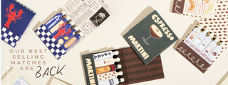 A styled flat-lay of multiple cocktail-themed matchbooks and small cards on a light beige background. Visible designs include a blue lobster checkered pattern, a dark green card reading 'ESPRESSO MARTINI', striped brown and white matchbooks, and a promotional text overlay that says 'OUR BEST SELLING MATCHES ARE BACK'.