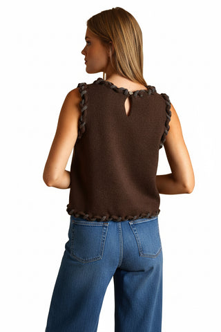 A woman shown from the back wearing a sleeveless brown knit dress with dark brown braided trim along the neckline, waist, and hem, set against a solid warm tan background.