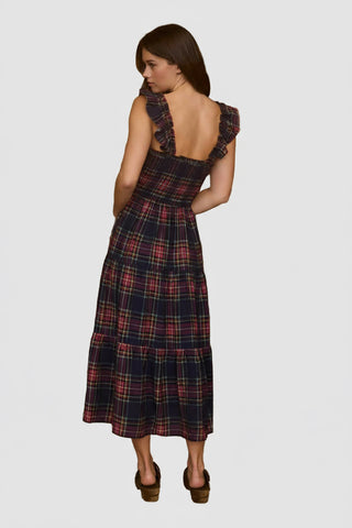 Woman in a plaid dress standing in front of a wooden wall with hats and a guitar.