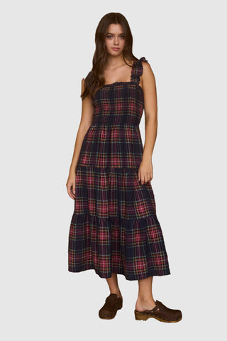 Woman in a plaid dress standing in front of a wooden wall with hats and a guitar.