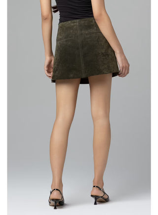 A model from the back wearing a dark olive green suede mini skirt, paired with a black top and black strappy kitten heel sandals, against a solid gray studio background.